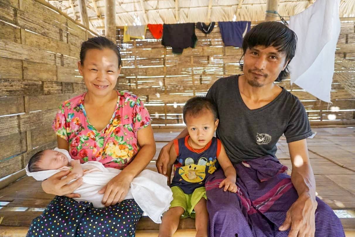 A father, a young boy and a mother holding a baby in a basic wooden home