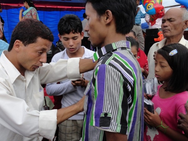 A man in a white shirt prays for a man in a multi-coloured shirt, while others pray in the background