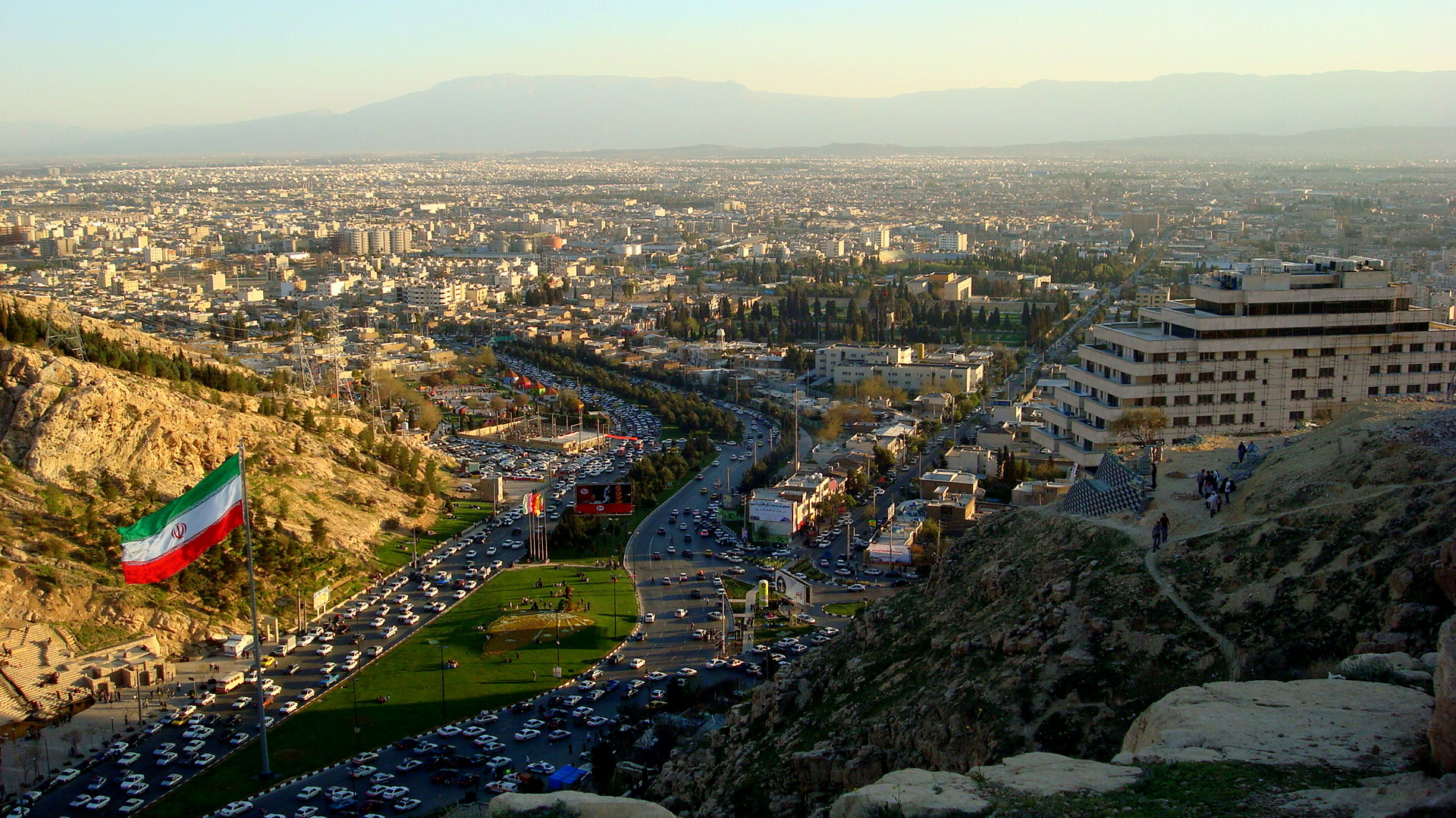 A city with mountains in the background and the flag of Iran in the foreground