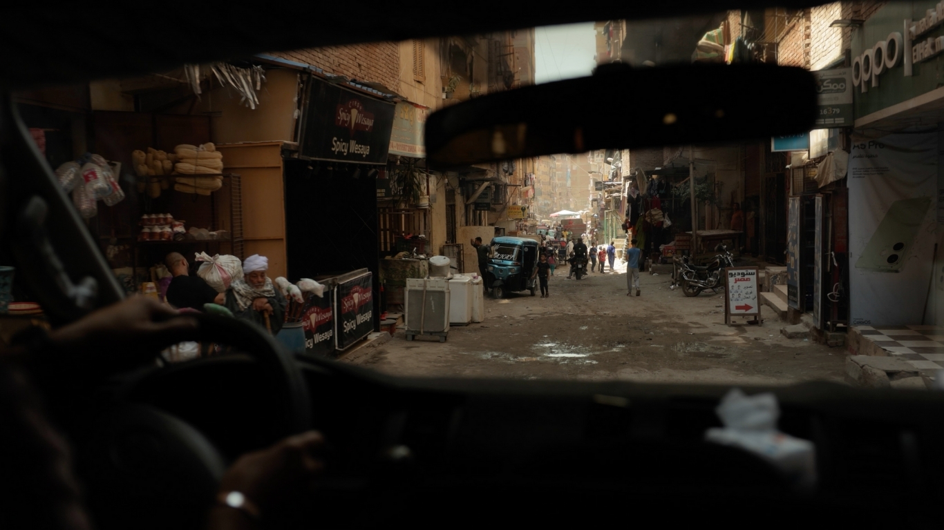 Inside a car, a narrow street in Egypt is visible, featuring close buildings and a glimpse of daily life.