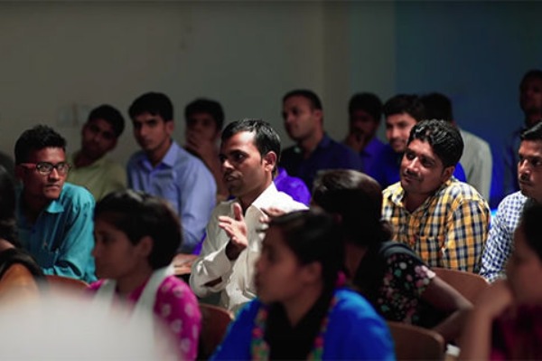 A man in a white shirt, sitting in the middle of a small audience of people, addressing the front of the room