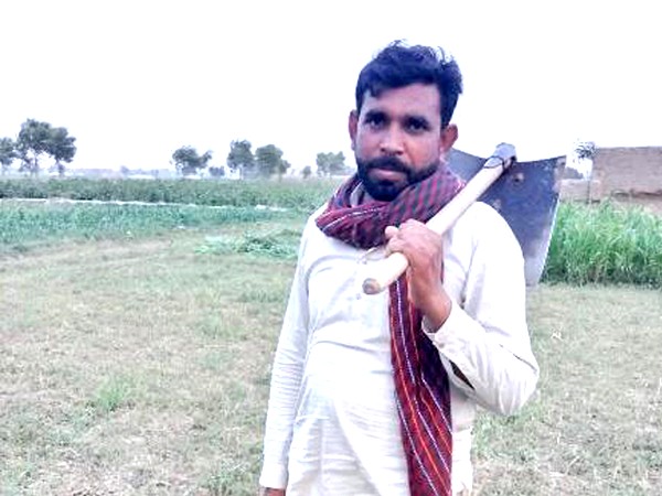 A middle aged man with dark hair and beard standing in a field holding a hoe