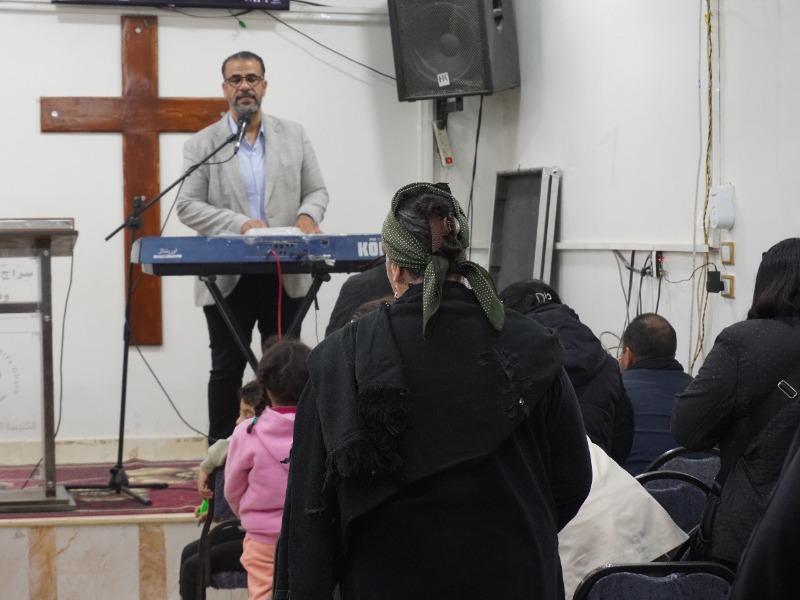 A man playing a keyboard at the front of a congregation with a cross in the background