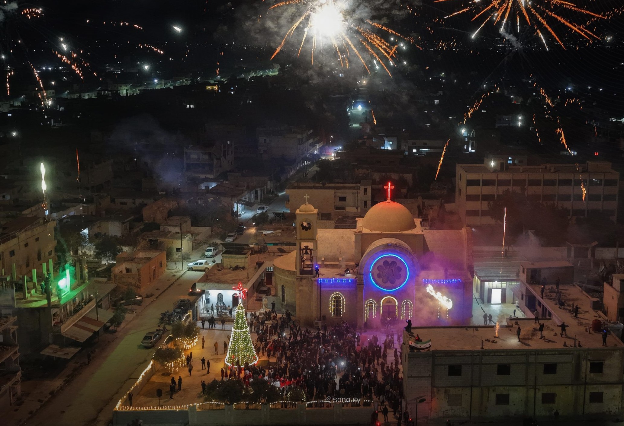 Fireworks above a church building with a red cross on the top and a Christmas tree at its side