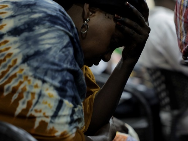 A woman in a blue patterned dress holds her hand to her head