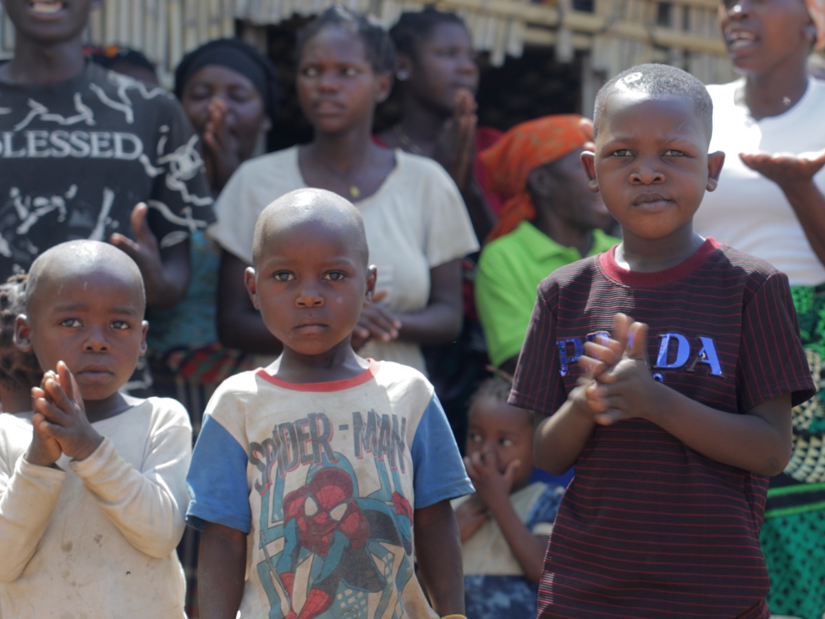 Three children standing at the front of a gathering outside a basic building