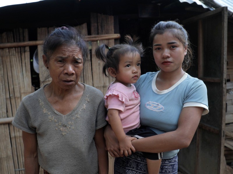 An older woman stands next to a younger woman holding a small girl, in front of a wooden house