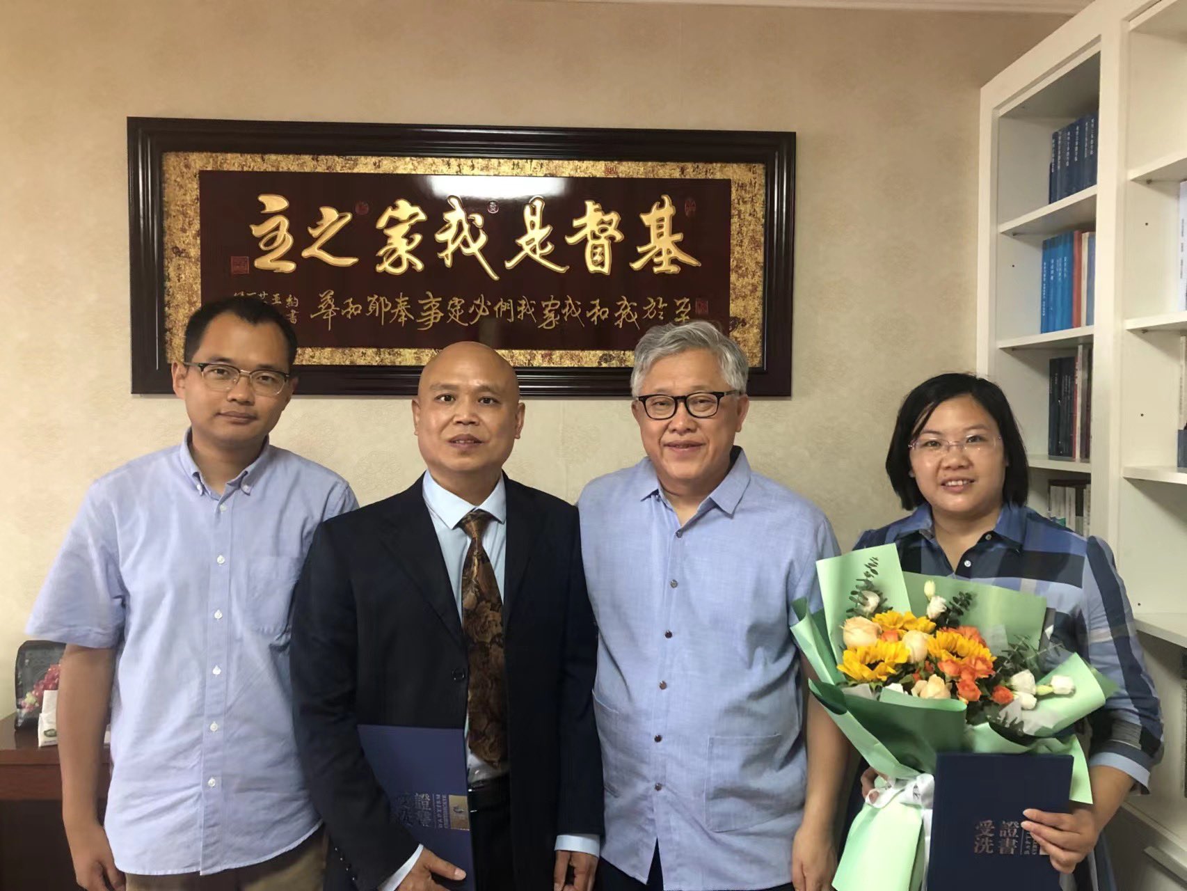 Man with grey hair and glasses wearing blue shirt, with a woman on his left holding flowers and two other men on his right