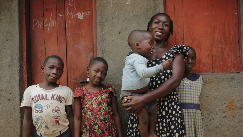 A Christian mother, Angela, stands with her children in front of a village building, capturing a warm family moment.