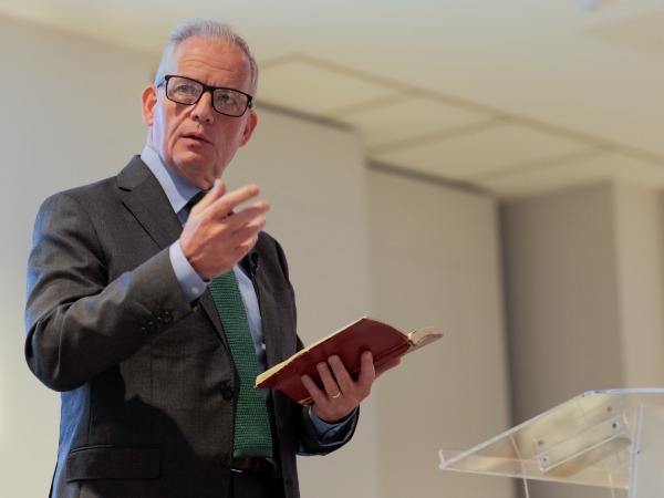 A man in a grey suit with green tie at lectern holding a Bible