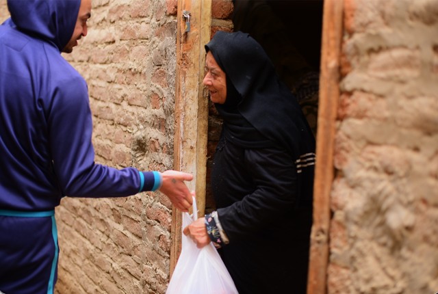 An elderly woman in black standing in a doorway receives a bag of food from a young man