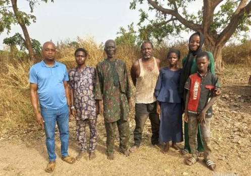 Four men, a woman and a boy, standing with a smiling man in front of some trees