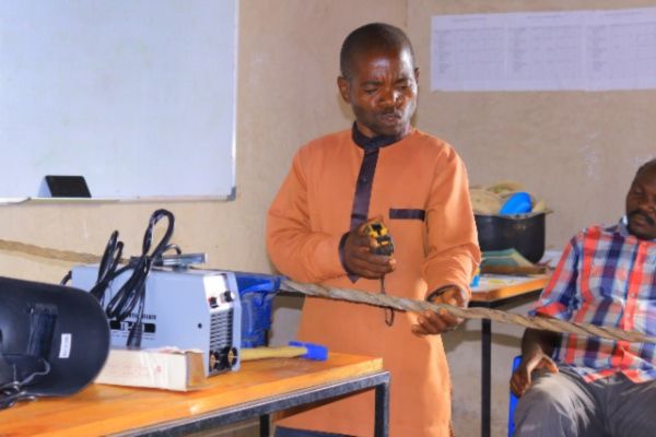 A man measures a metal cable in a classroom, illustrating hands-on learning supported by Barnabas Aid&#039;s project partners.